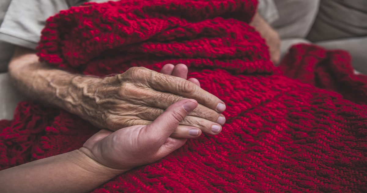 An elderly individual rests their hand on a loved ones hand on a red blanket