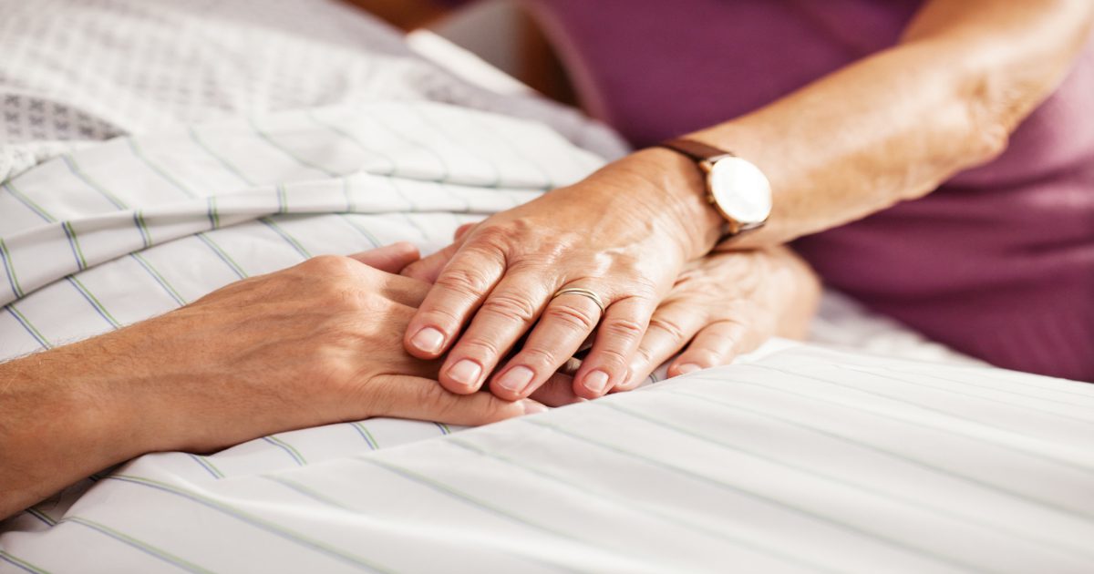 An elderly man lays in bed while an elderly woman places her hand on his for comfort